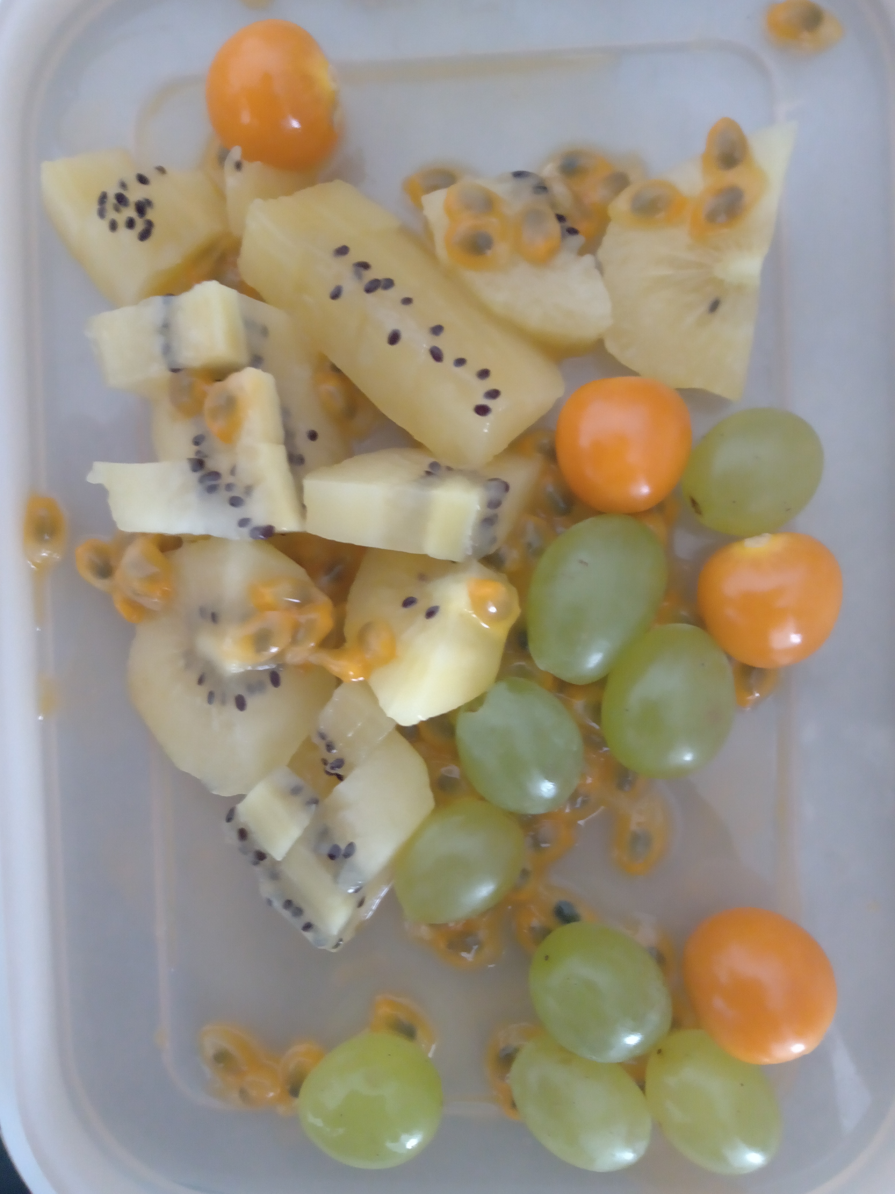 Na zdjęciu widać kompozycje z owoców ułożoną na plastikowej, przezroczystej, prostokątnej pokrywce. Widzimy:pokrojone w cząstki kiwi, zielone winogrona i owoce miechunki.