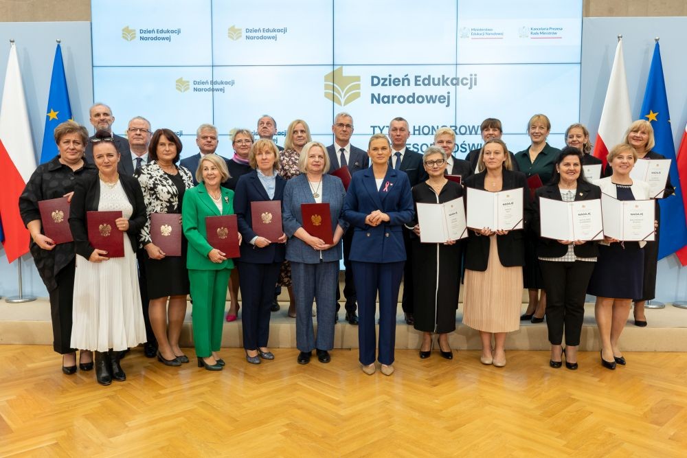 Grupa nauczycielek i nauczycieli pozuje do zdjdjęcia, trzemając dyplomy. W tle ściana z napisem Dzień Edukacji Narodowej.